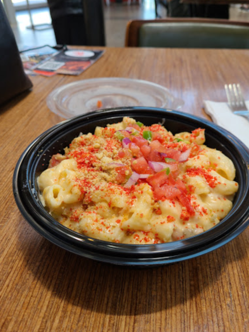 A bowl of macaroni and cheese topped with diced tomatoes, onions, and crushed red seasoning on a wooden table.